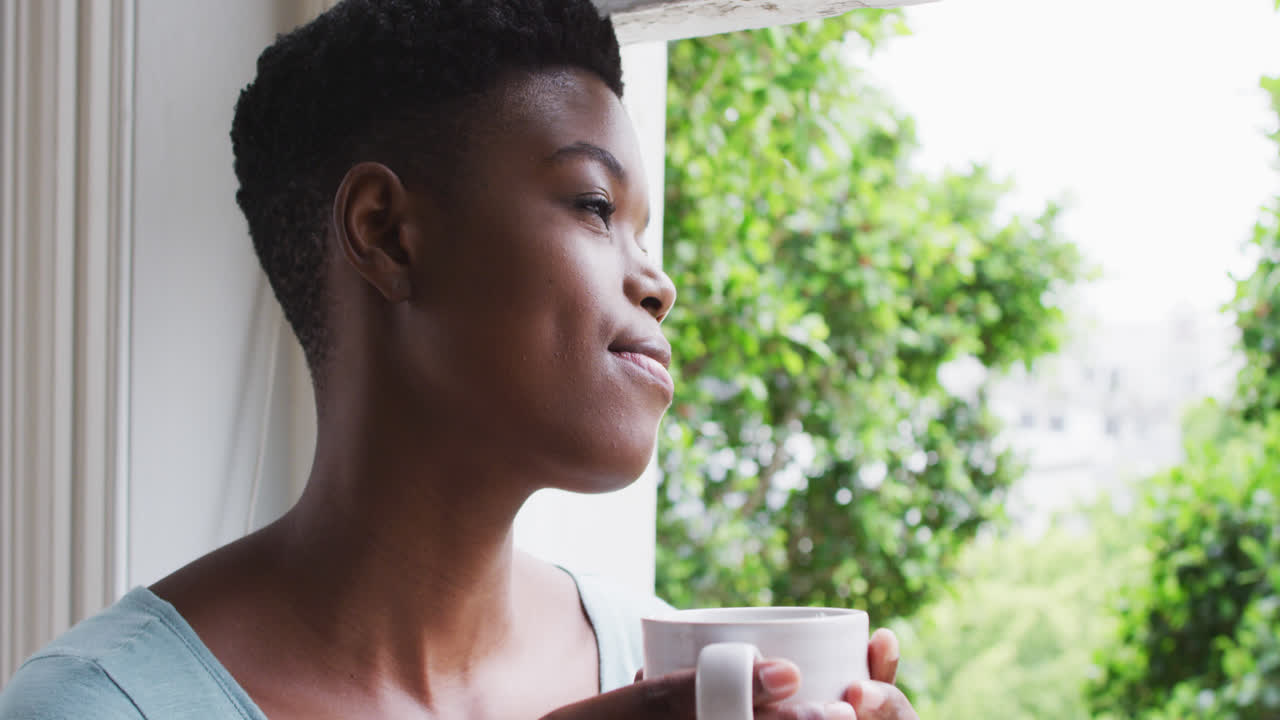 African american woman holding coffee cup looking out of window at home