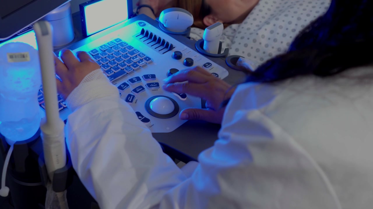 Primo piano delle mani di uno specialista medico che utilizza la tastiera e la trackball di un ecografo durante l'esame di un paziente in una stanza buia con illuminazione blu