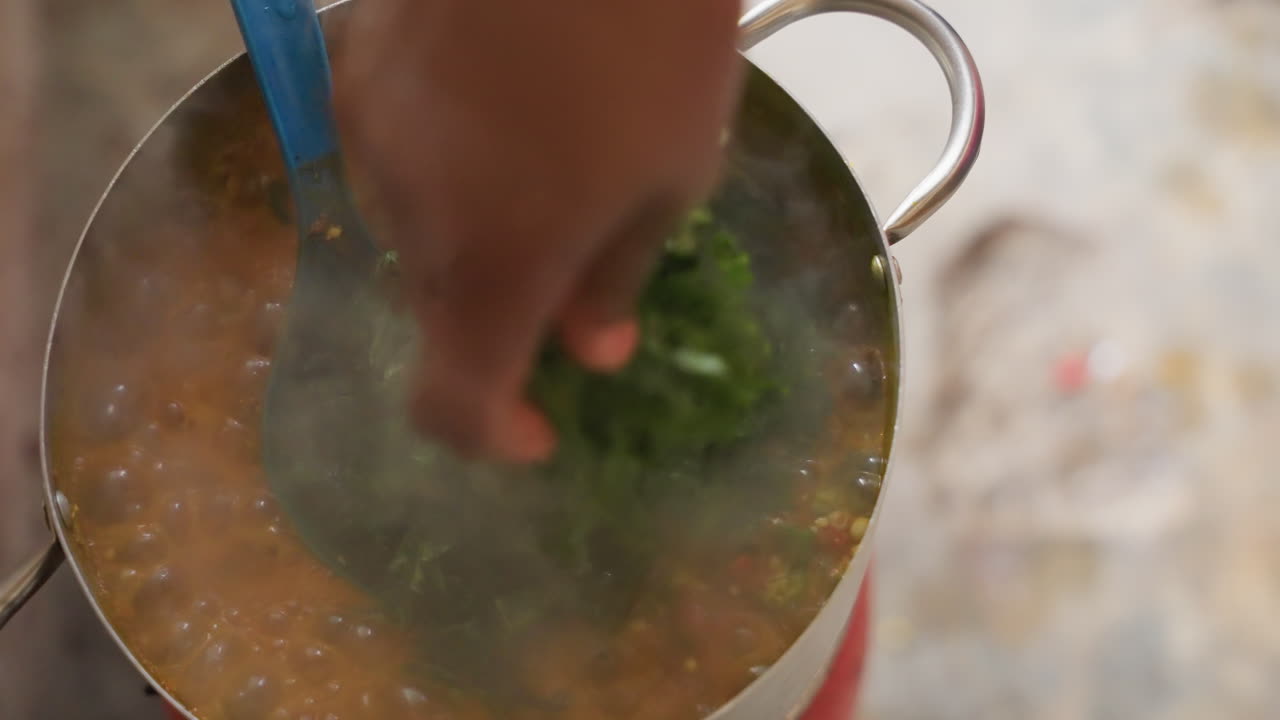 Cuoco nero che mescola una pentola con verdure, aggiungendo erbe tritate da una ciotola rosa in uno stufato di pomodoro e lenticchie, un mestolo blu mescola il vapore che sale, bancone rustico, primo piano di mani che preparano un sostanzioso pasto in famiglia.