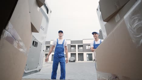 Two young workers of removal company unload boxes and furniture from minibus into customer's home