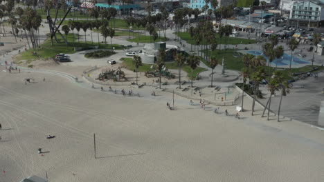 AERIAL:-Flight-towards-Venice-Beach-skatepark-and-boardwalk-with-palm-trees-and-bike-lane,-Sunny,-Los-Angeles-California