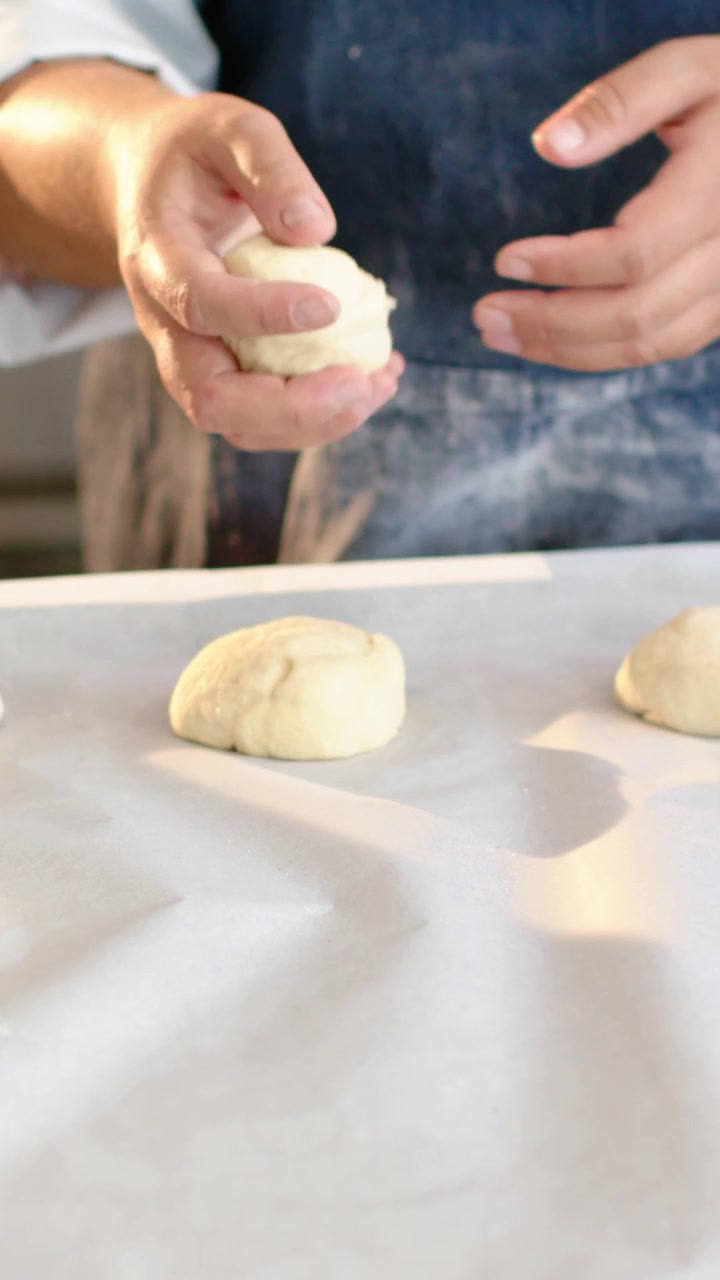 Vertical video: Reaching down denim-aproned baker shaping dough in kitchen, using parchment to bake
