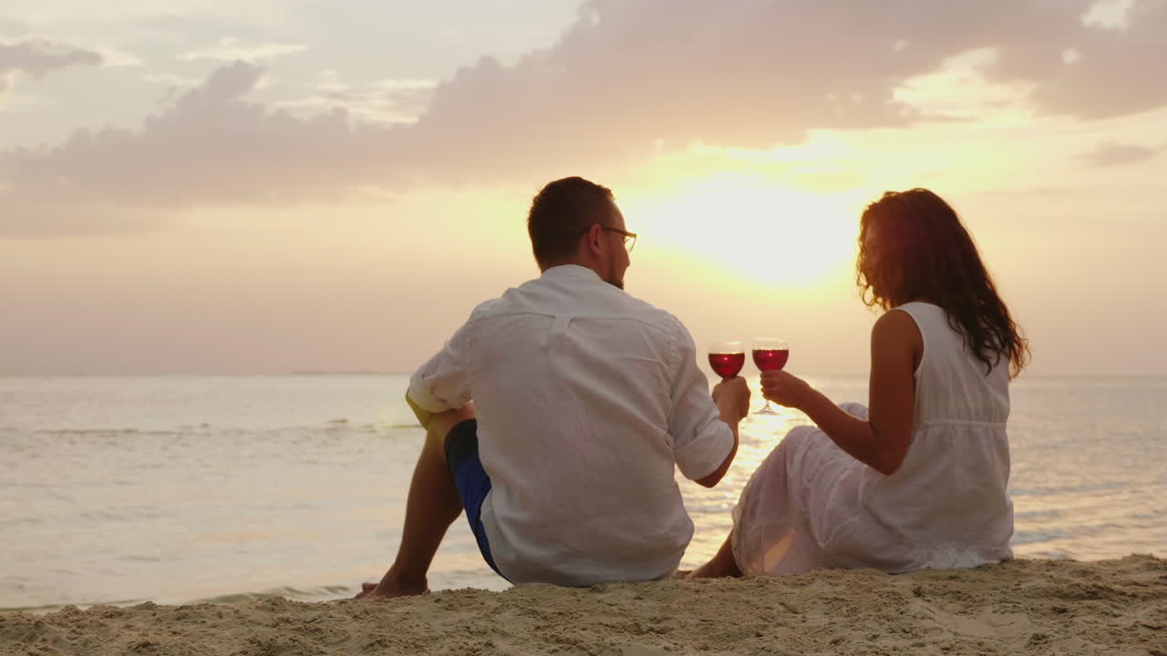 Una giovane coppia beve vino in riva al mare al tramonto. Sono seduti sulla sabbia, fanno tintinnare i calici - Anniversario