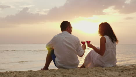 a young couple is drinking wine by the sea at sunset they sit on the sand clink glasses anniversary