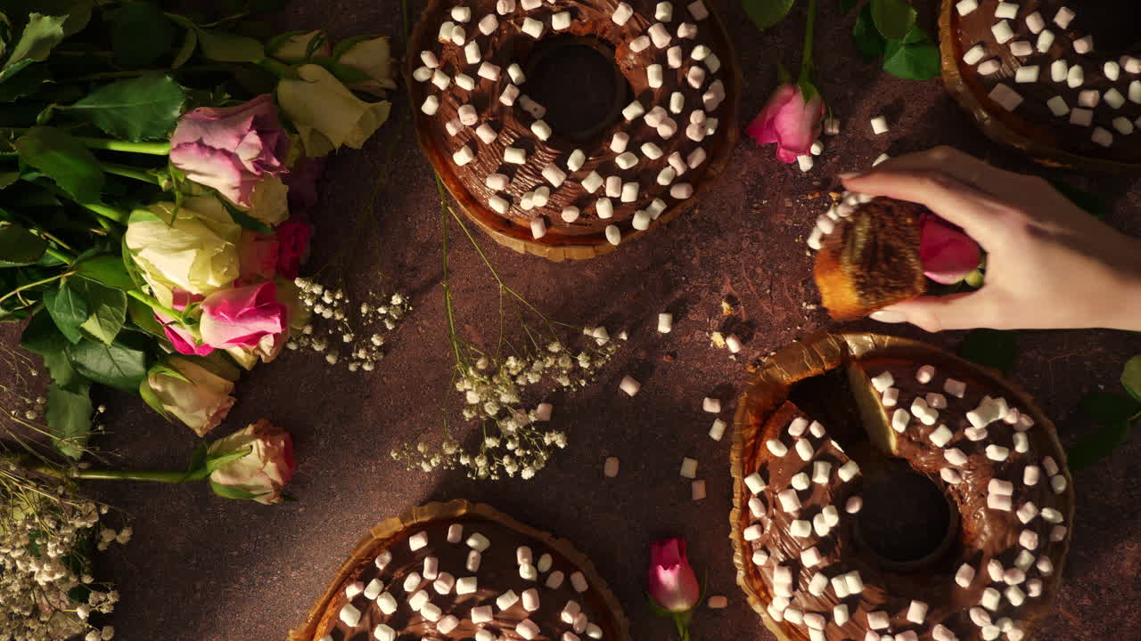 Vista dall'alto di una mano che prende una fetta da una torta al cioccolato a forma di ciambella guarnita con mini marshmallow. Rose decorative e dolcetti creano un romantico tavolo da dessert per compleanni e San Valentino