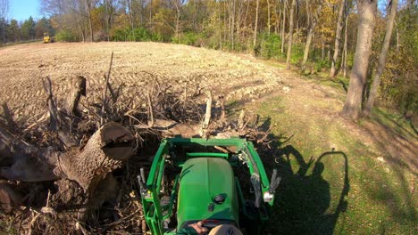 punto de vista de ángulo alto en un pequeño tractor verde usando horquillas elevadoras para recoger un tocón de árbol de un montón y mover escombros cerca de los bosques a principios de otoño