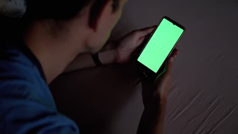 hombre en la oscuridad en la cama sostiene y examina el teléfono inteligente con pantalla táctil verde