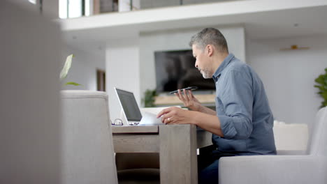 Hombre-Birracial-Mayor-Usando-Un-Teléfono-Inteligente-Y-Una-Computadora-Portátil-En-El-Escritorio,-Trabajando-Desde-Casa,-Copiando-Espacio,-Cámara-Lenta