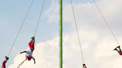 Papantla-Flyer,-Die-An-Den-Knöcheln-Mit-Einem-Seil-Festgebunden-Sind,-Das-Um-Eine-Metallstange-Gewickelt-Ist