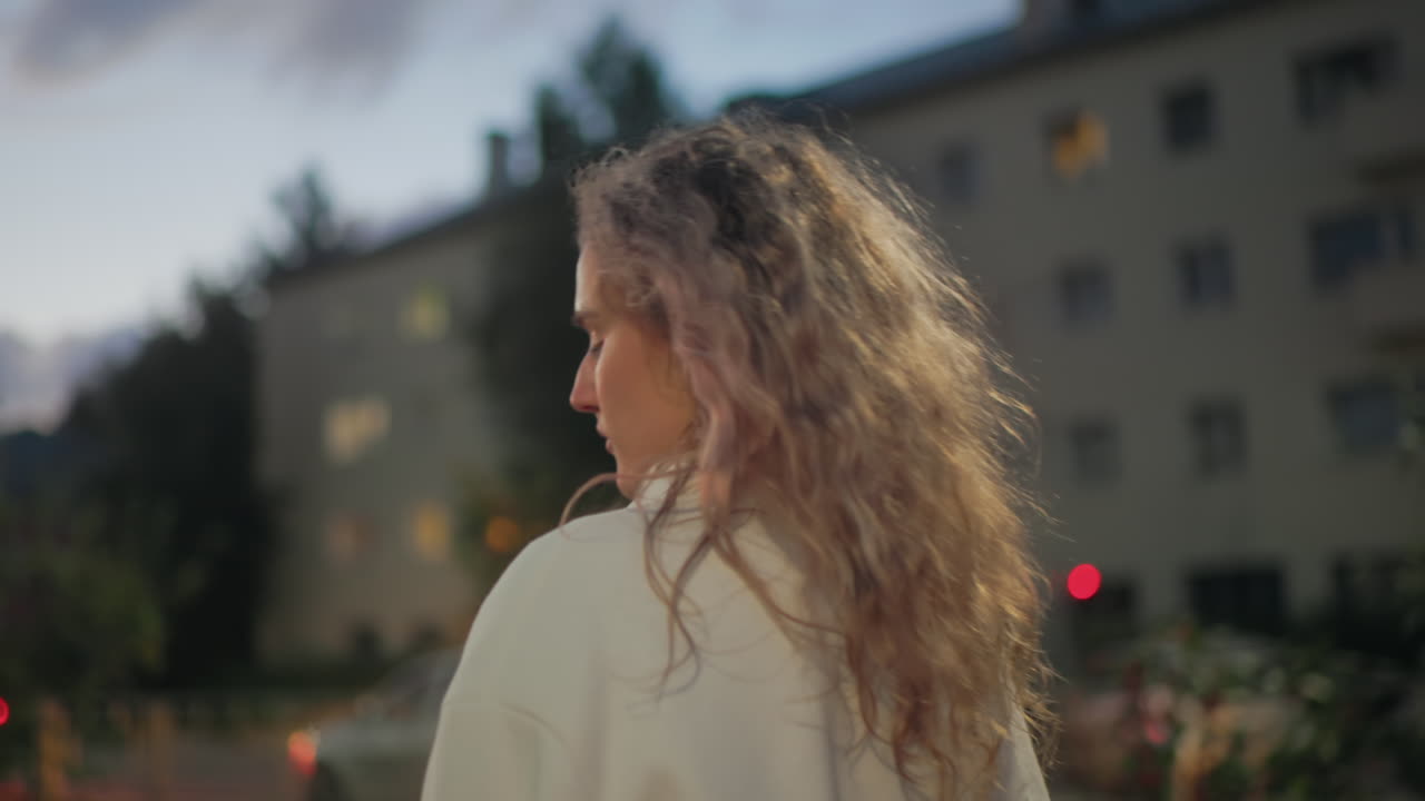 Side view of pensive woman with curly hair holding umbrella walking past residential buildings with blurred glowing lights and parked vehicles during quiet evening in soft urban atmosphere