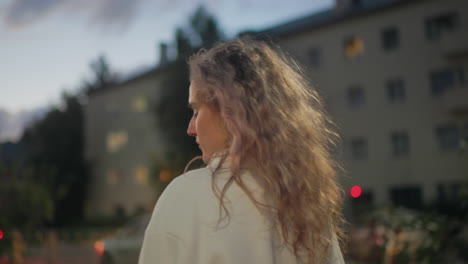 side view of pensive woman with curly hair holding umbrella walking past residential buildings with blurred glowing lights and parked vehicles during quiet evening in soft urban atmosphere