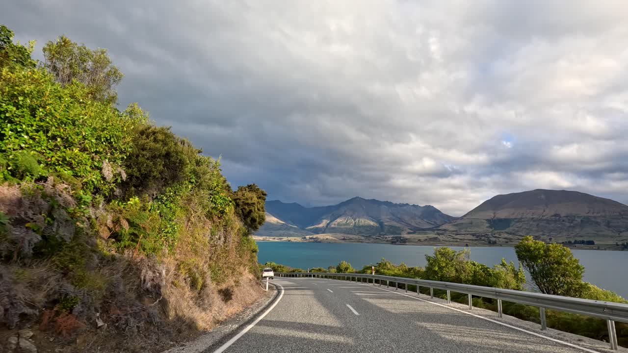 Vehicle travels winding lakeside road, revealing dramatic mountains, cloudy sky, and lush greenery