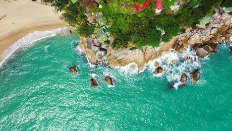 Waves-washing-on-the-rocky-shore-with-secluded-sandy-beach,-Aerial-overhead-of-the-tropical-island-coast