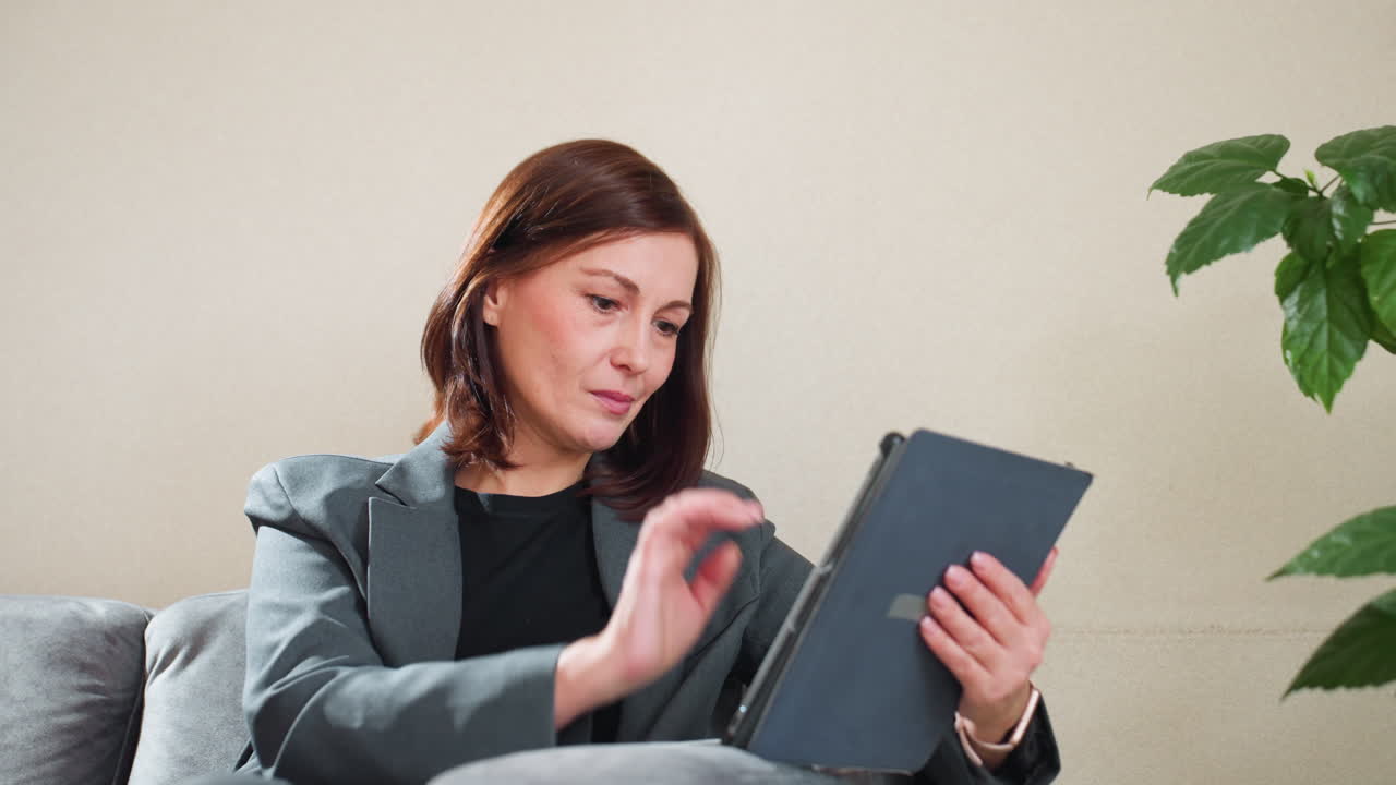 Confident businesswoman in gray blazer using digital tablet while sitting on sofa in bright modern office space with plant in background, engaged in online communication or work-related task