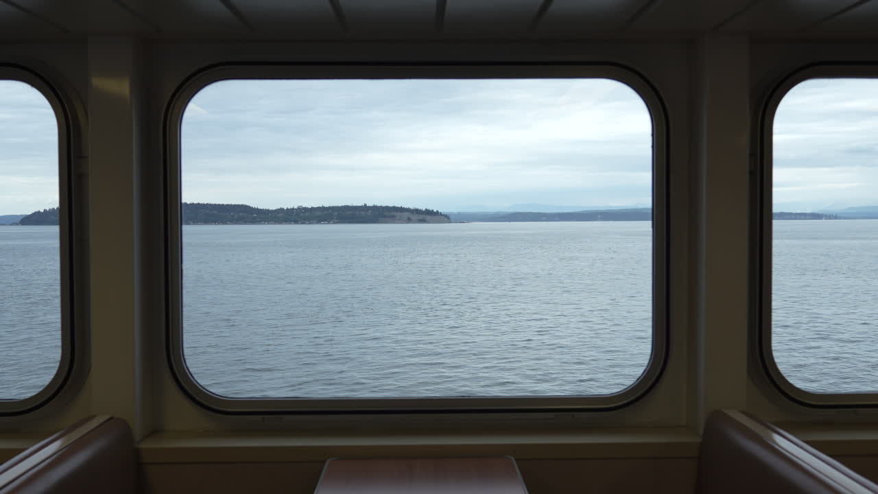 Videoaufnahme einer friedlichen Szene aus dem Inneren einer Fähre, mit Blick durch große Fenster auf das ruhige Wasser und die ferne Küstenlinie unter einem bewölkten Himmel