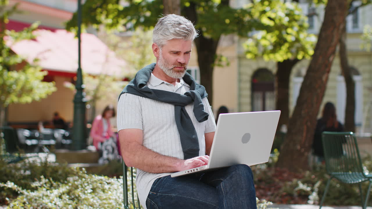 Middle-aged man opens laptop start working sends messages sitting on chair in urban city street