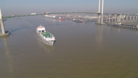 aerial tracking shot of a cargo ferry passing under the qe2 dartford bridge on the river thames that links essex to kent in the uk