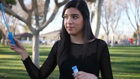 attractive hispanic woman blowing bubbles and smiling with joy and happiness as she has fun playing in the sunny park