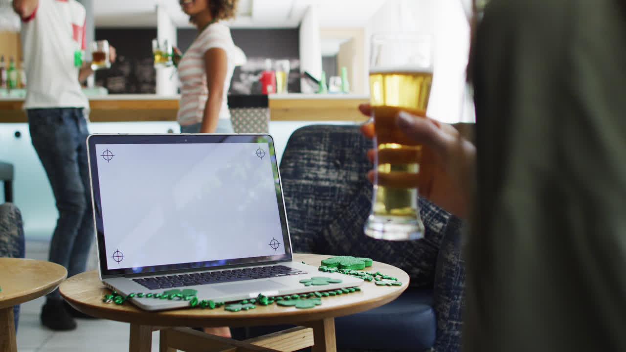 Mixed race woman celebrating st patrick's day making video call with laptop at a bar