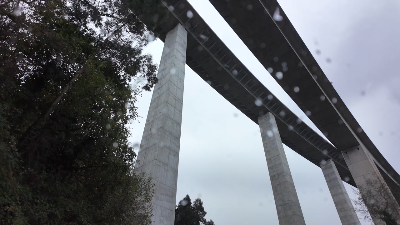 Car passes beneath a large concrete highway bridge with dramatic clouds overhead, captured in slow motion.