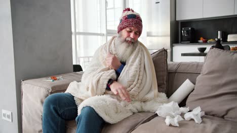 Portrait-of-a-sick-elderly-man-with-a-lush-beard-and-a-red-hat-wrapped-in-a-white-blanket-and-folds-used-napkins-near-him-while-sitting-on-the-sofa-near-a-thermometer-and-a-set-of-medications-in-a-modern-apartment-on-the-sofa