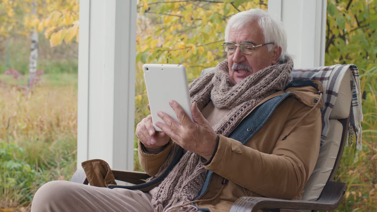 Eldre mann sitter på en benk i hagen hjemme mens han snakker i en videosamtale ved hjelp av et nettbrett.