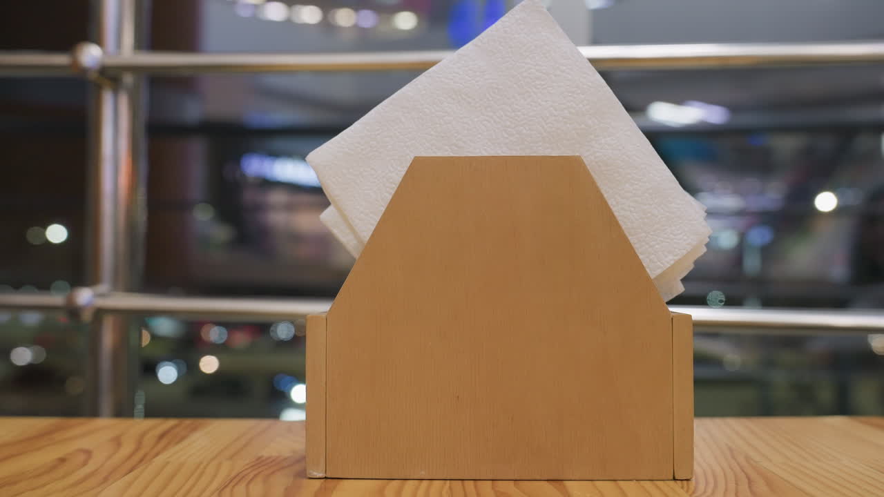 Close up wooden napkin holder with neatly folded white tissues placed on restaurant table under soft evening lighting with blurry city lights glowing through window in background