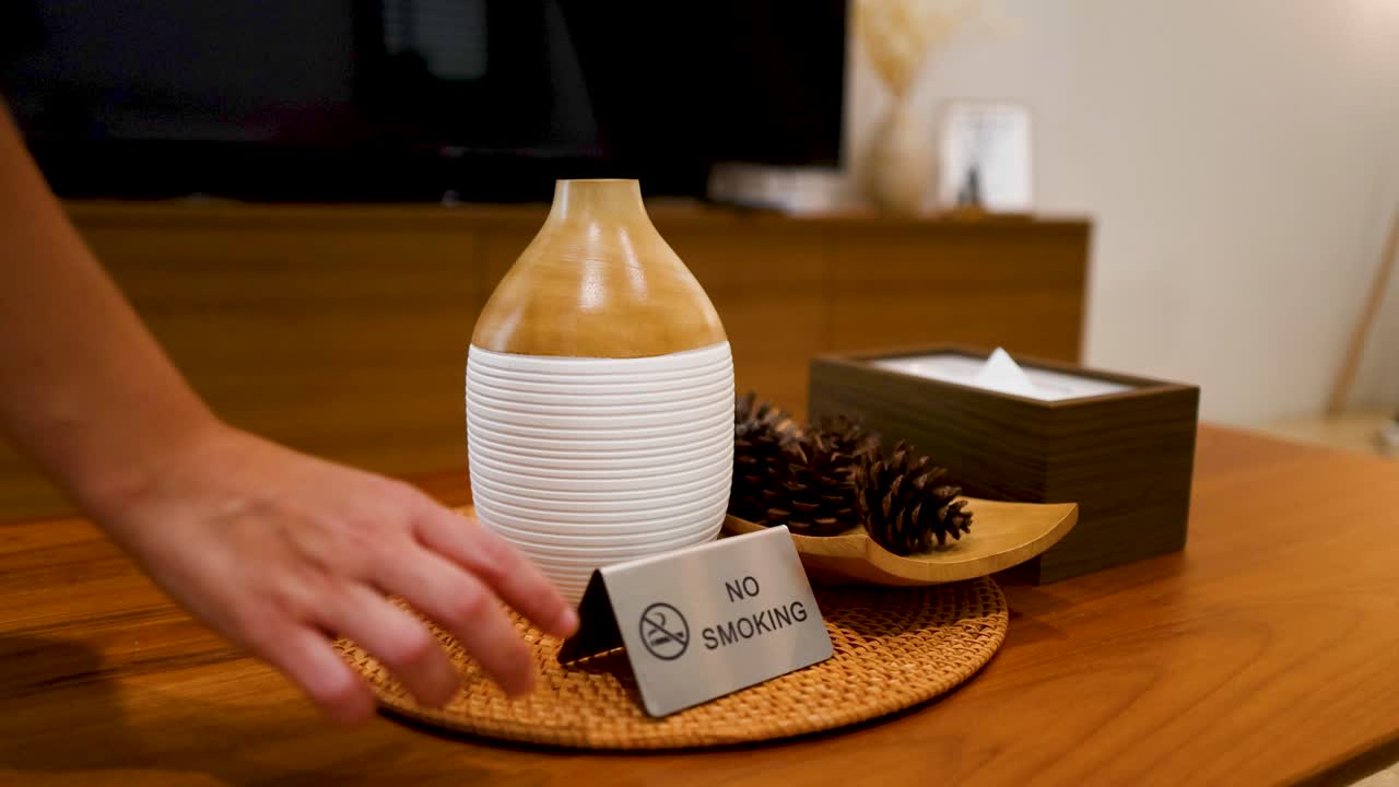 Une main plaçant un panneau métallique "interdit de fumer" sur une table en bois à côté d'un diffuseur d'aromathérapie.