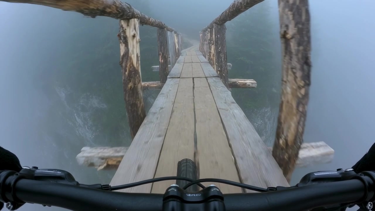 Avvicinandosi a un ponte, un ciclista adulto pedala lungo uno stretto ponte di assi per raggiungere l'altra sponda, con il manubrio.