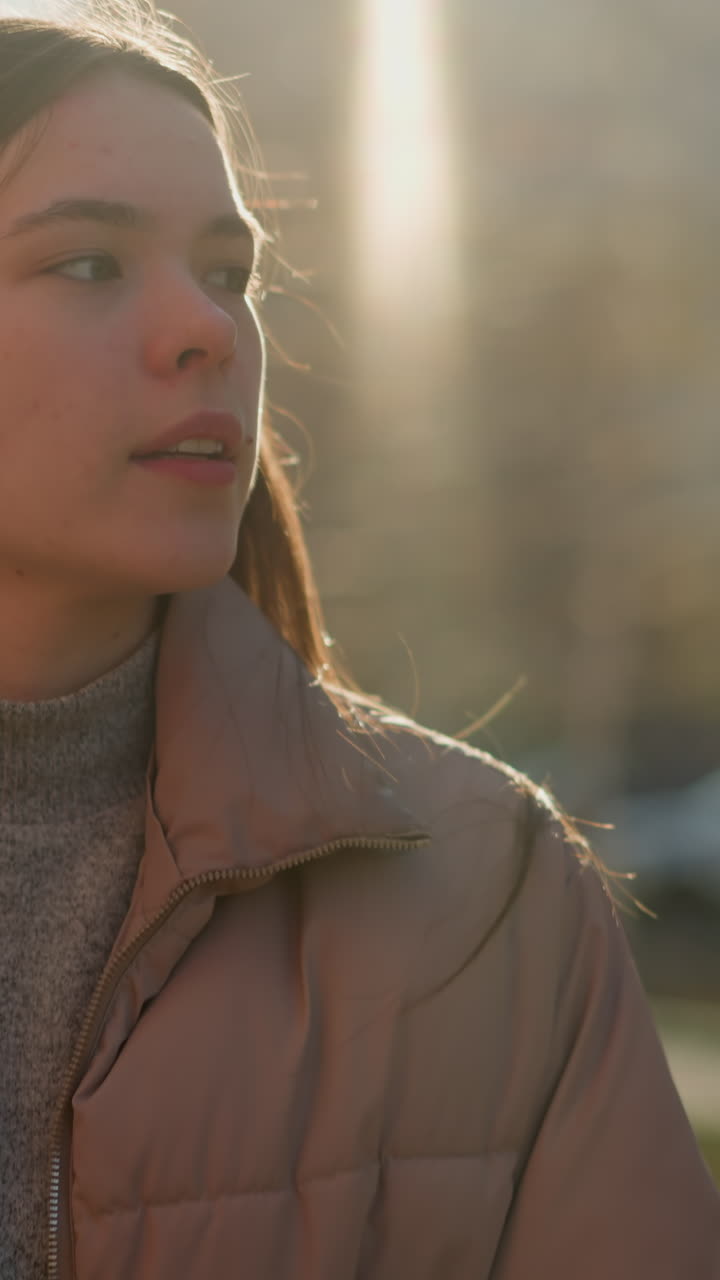 Front view of a girl walking through an urban area at sunset, wearing a peach jacket and a grey inner shirt. The sunlight softly illuminates her face as she gazes ahead with a calm expression