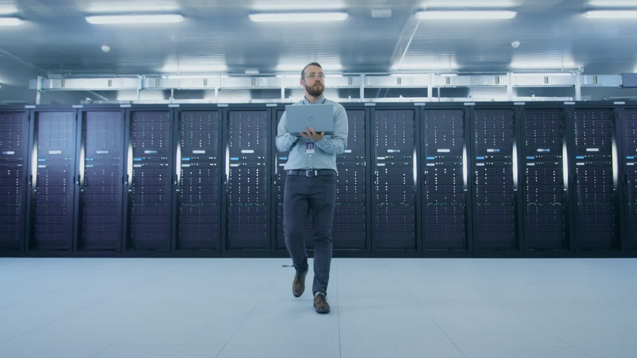 el centro de datos barbudo es un profesional que se aleja del corredor de los gabinetes de servidores de trabajo con una computadora portátil. lleva pantalones vaqueros y camisa grises.