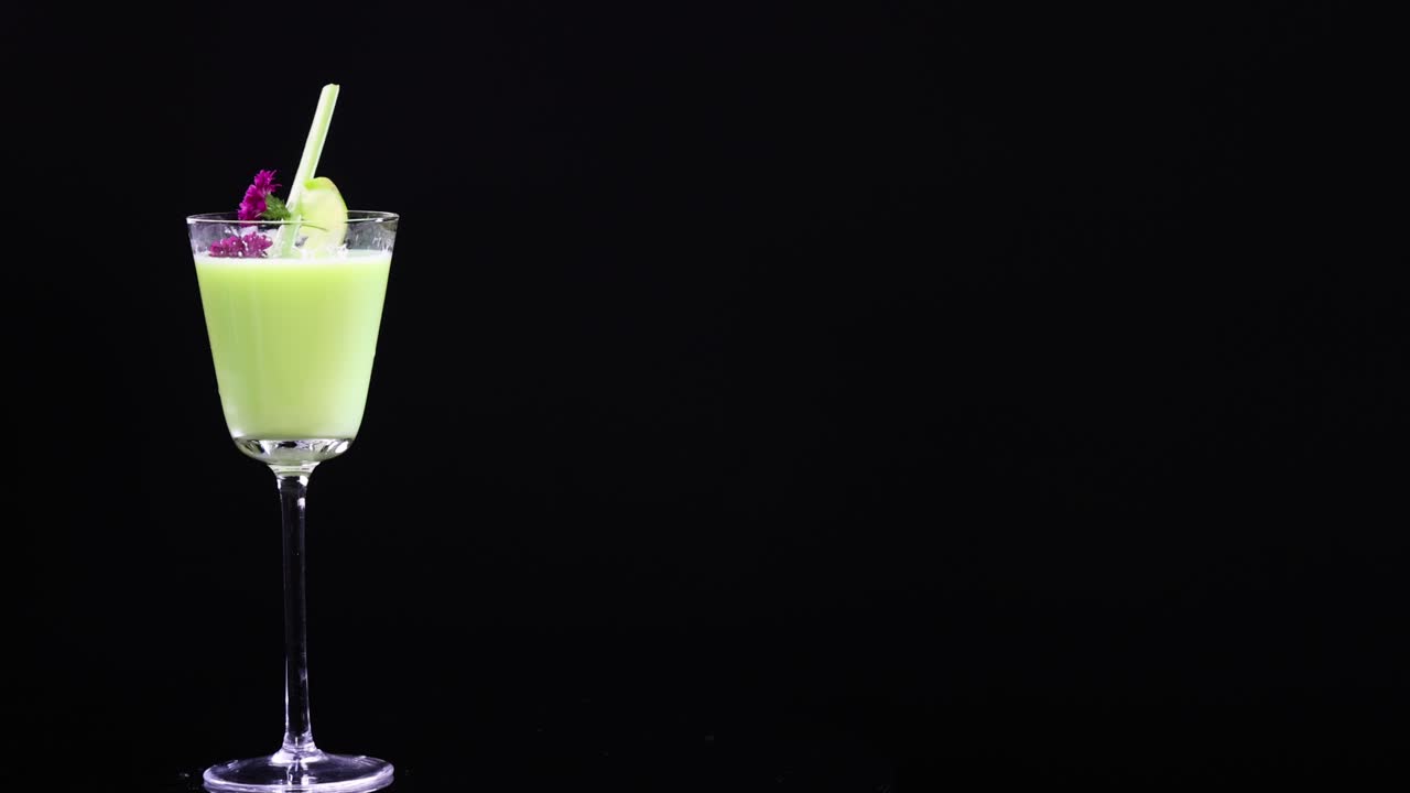 A vibrant green cocktail in a glass with lime and flower garnish against a black background