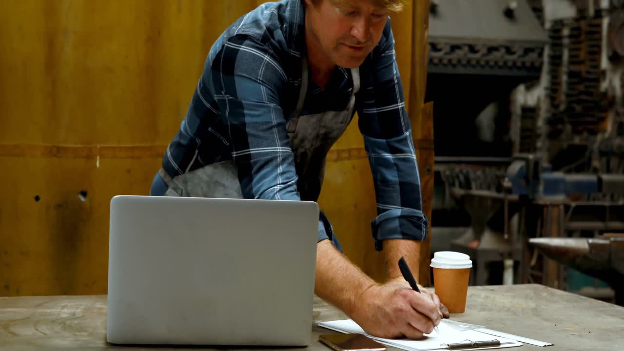 Blacksmith writing on clipboard while using laptop 4k