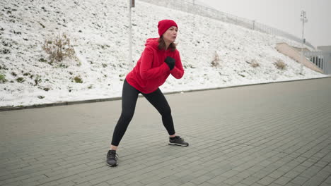 side view of woman performing outdoor workout in winter, stretching into a side lunge on paved ground near snowy hill and urban landscape, she wears a red hoodie and black leggings