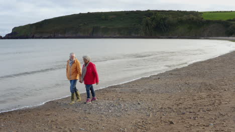 drone shot of senior couple holding hands as they walk along shoreline on winter beach vacation