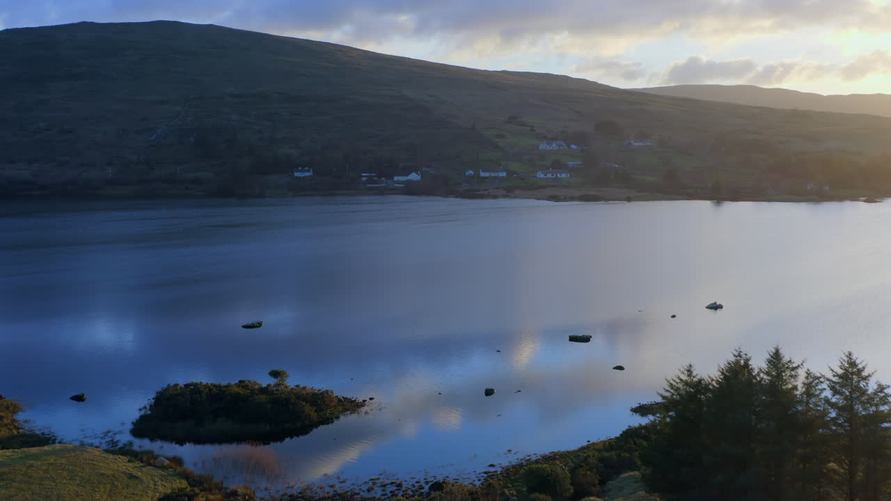 Aerial pullback of glistening orange glow on clouds reflecting on Lough Bofin at dusk, where soft pink and blue hues reflect on the water’s surface, Connemara, Galway, Ireland