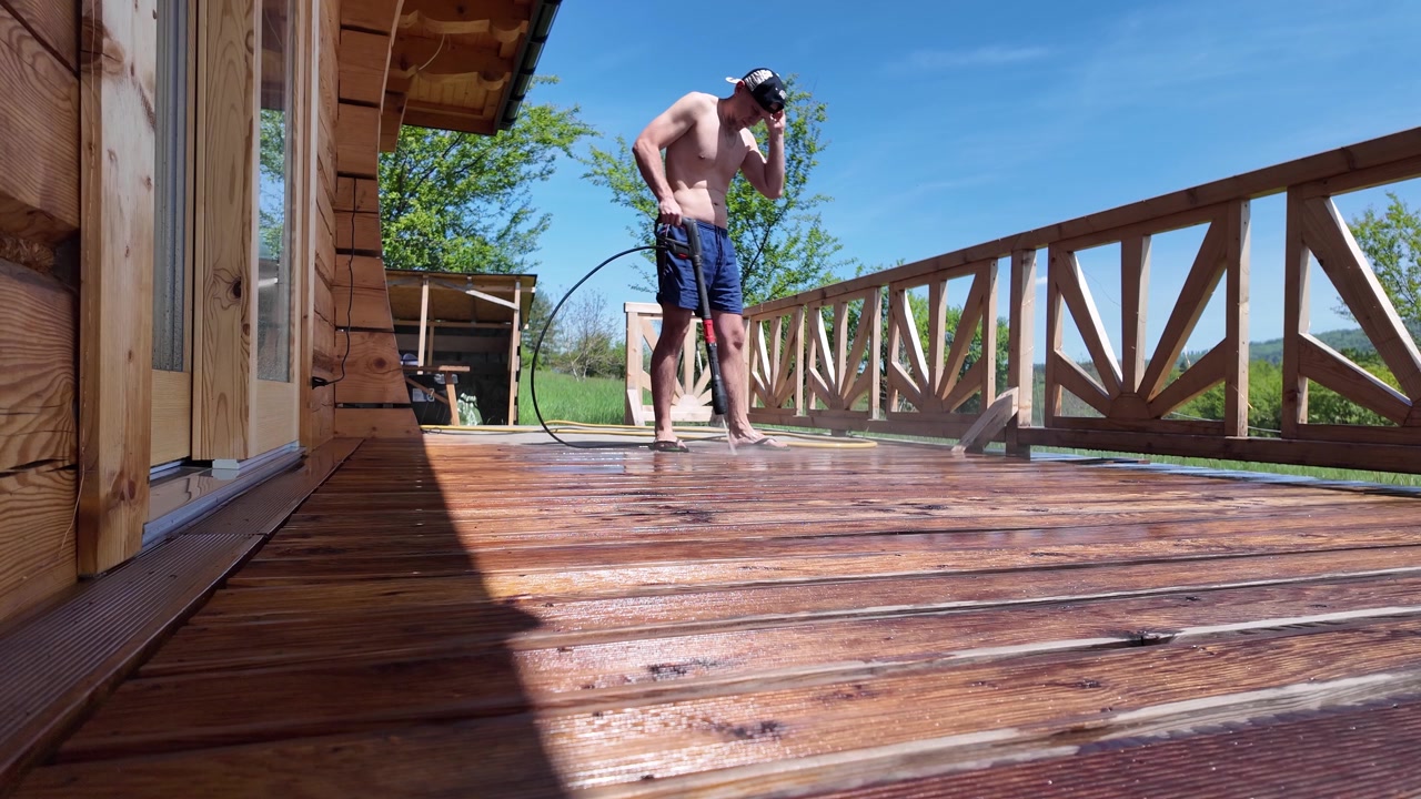 Vista dal basso di un uomo in pantaloncini e berretto da baseball che usa un'idropulitrice per lavare una terrazza in legno presso una baita tradizionale in estate