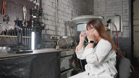 female technician in white coat picks up protective face shield, puts it on, and relaxes back on seat in auto repair shop with light, machinery, and equipment in background