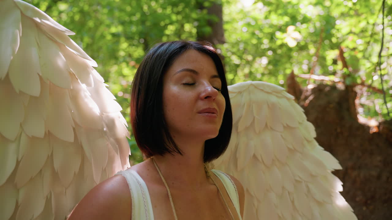 Primo piano di una donna in costume da fata con grandi ali bianche in piedi nella foresta, occhi chiusi in meditazione pacifica, luce del sole che filtra attraverso le foglie verdi, che esprime serenità