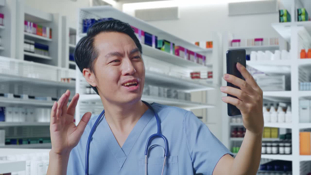 스테토스코프를 들고 있는 아시아 남성 의사가 손을 흔들고 약국에 서서 스마트폰으로 비디오 통화를 하고 있다.