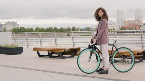 hermosa mujer rizada con ropa formal mirando y sonriendo a la cámara mientras se sienta en una bicicleta en el puente de la ciudad 2