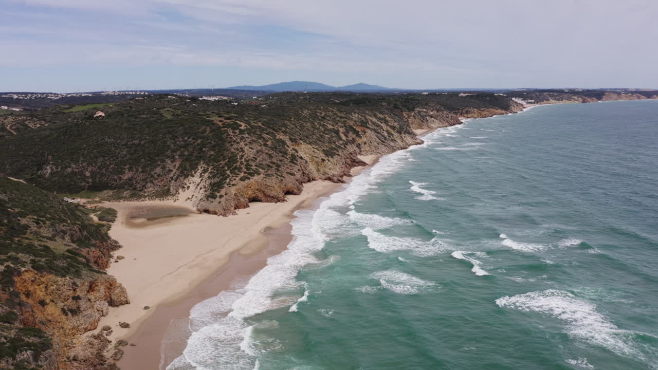Aerial of wild stormy ocean waves under cloudy sky and sandy beach, with cliffs along coastline