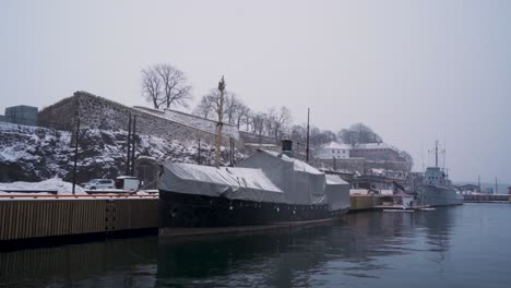 covered boat and navel vessel moored at harbour beside akershus castle during winter