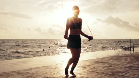 Young-woman-working-out-on-the-jump-rope-against-the-son-by-the-beach-in-Slow-Motion.-Girl-jumping-on-a-skipping-rope-by-the-sea