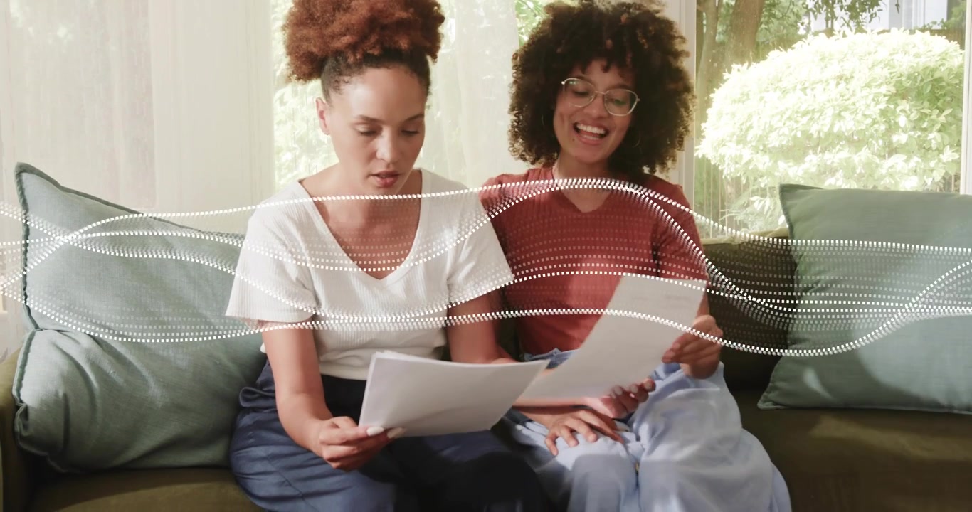 Vrouw van middelbare leeftijd leest financiële krant hardop voor terwijl vriendin voorover buigt, wijst en secties vergelijkt.
