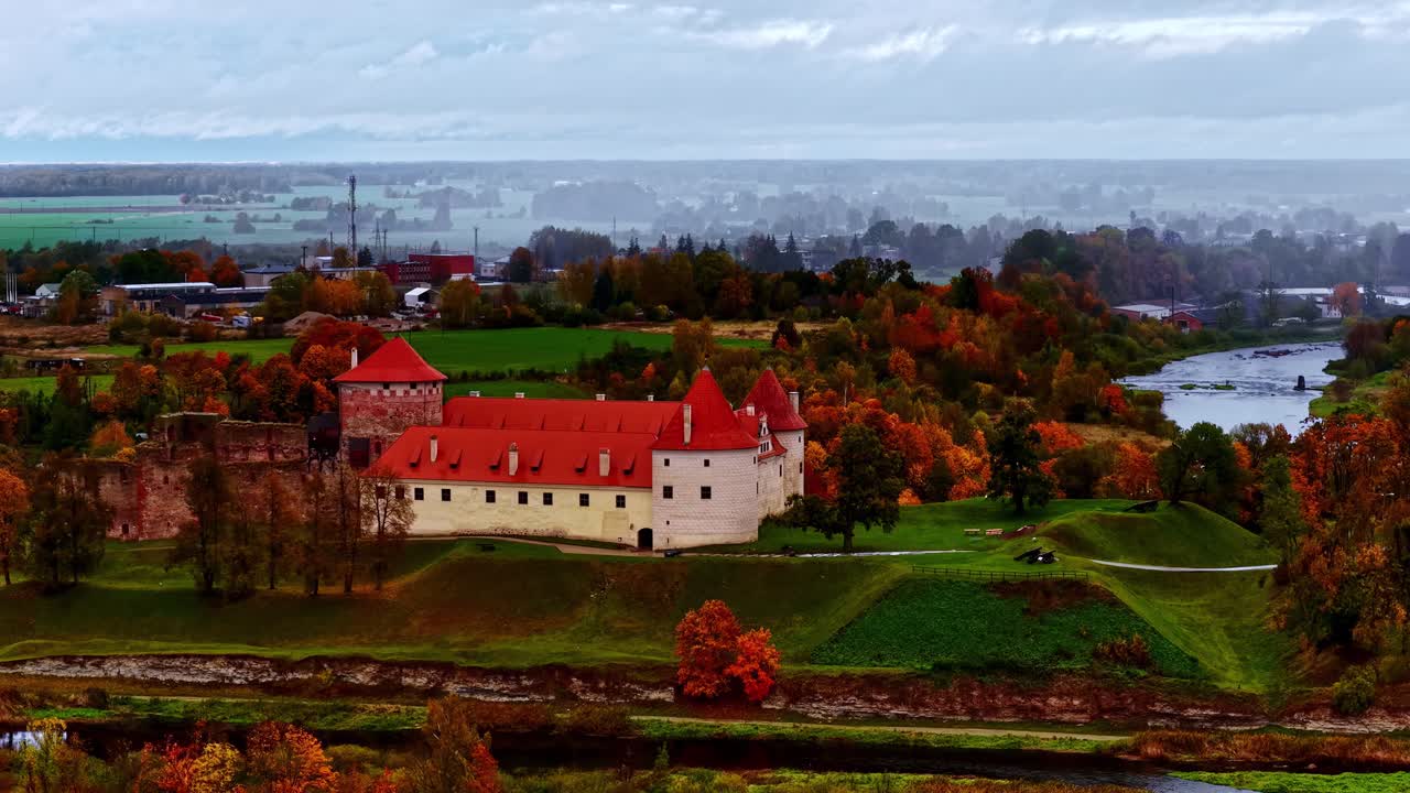 Aerial of castle with red roofed buildings surrounded by fields and trees, looking over farmland
