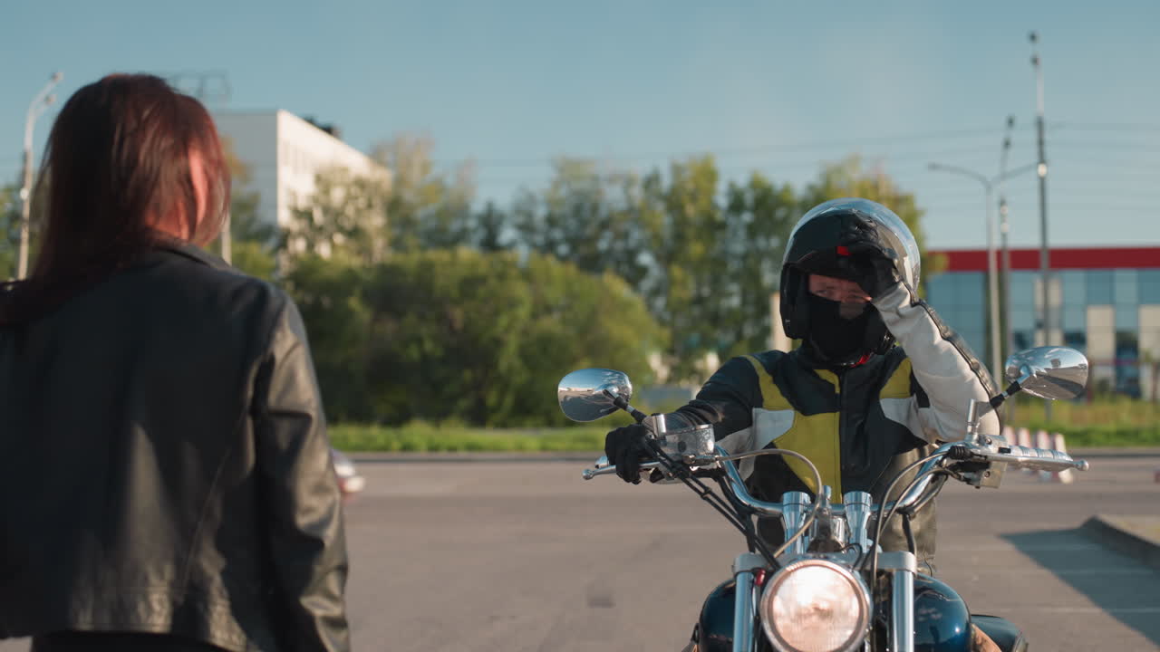 Woman in leather jacket extends arm to stop motorcycle as rider approaches and opens helmet under bright daylight on urban street with buildings and traffic in background