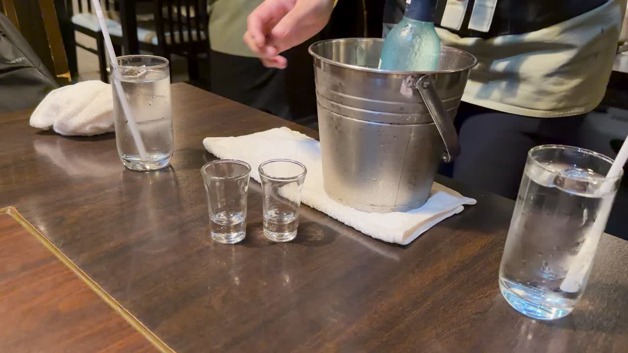 Server pours cold sake from icy bottle into shot glasses on wooden table, natural lighting