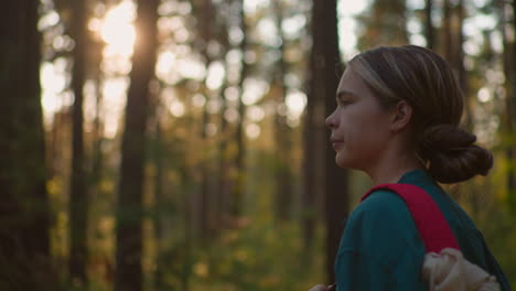 side view of tired hiker in green shirt holding red bag while walking through sunlit forest, warm sunlight filtering softly through trees, casting an ethereal glow on her as she gazes downward