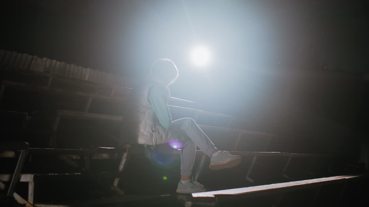 Trainer sitting on empty stadium bleachers at night holding bulldog lovingly on her legs under powerful spotlight, creating touching moment of loyalty, care, and quiet emotional bond in dark setting